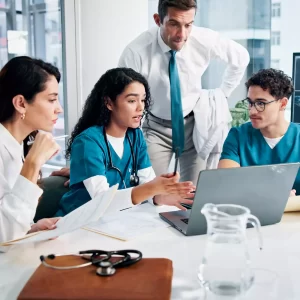 A diverse group of four medical professionals, including doctors and nurses, gathered around a laptop in an office, discussing information and collaborating. A stethoscope is on the desk in the foreground.