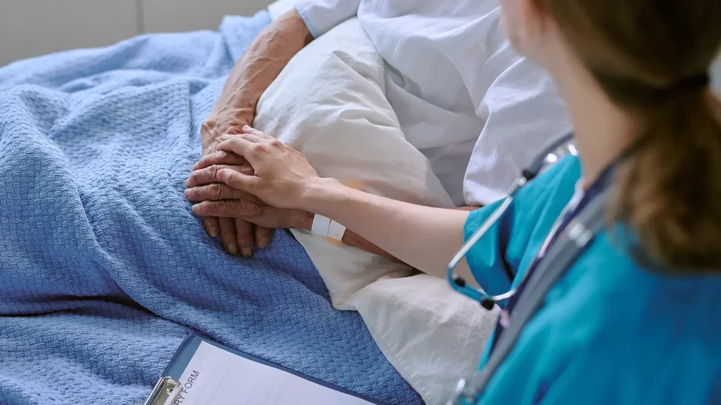Senior Caucasian patient lying in hospital bed receiving comforting touch from young adult Caucasian female nurse